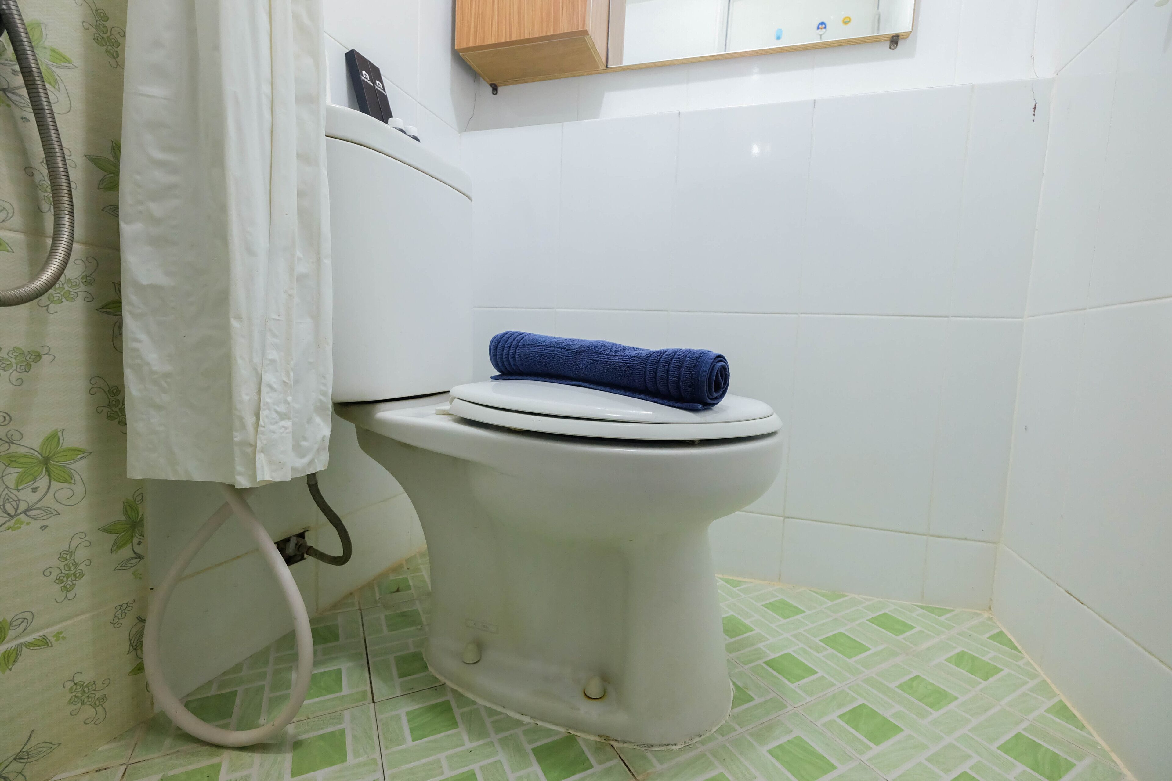 A white toilet with a blue towel on the seat, a shower hose, and a tiled floor with a green pattern.
