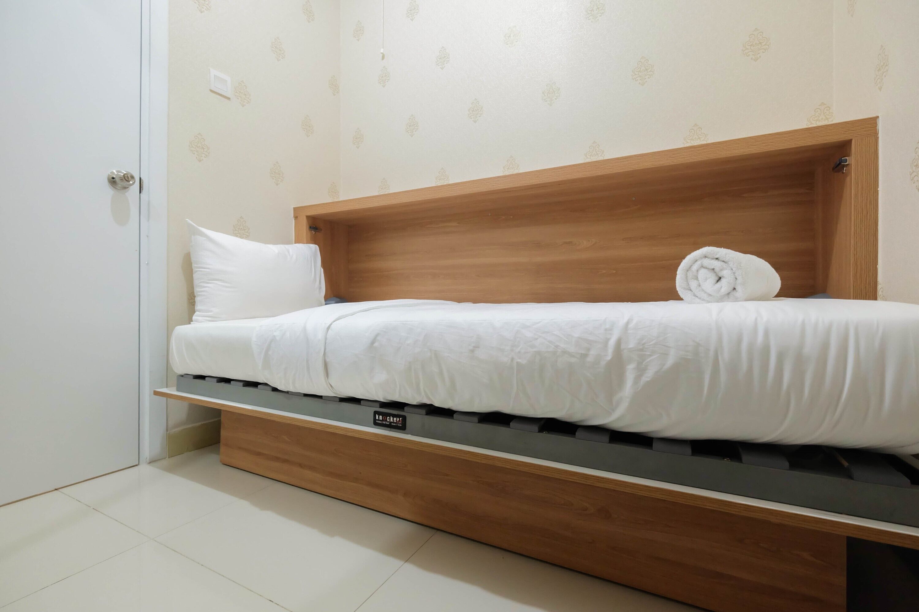 Una cama compacta con cabecera de madera, almohada blanca y una toalla enrollada cuidadosamente.