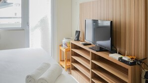 A hotel room with a bed, a TV on a wooden stand, a small table with a coffee maker, and a window with a view of buildings. - Cozy Studio Green Pramuka Apartment next to Mall (Jakarta)