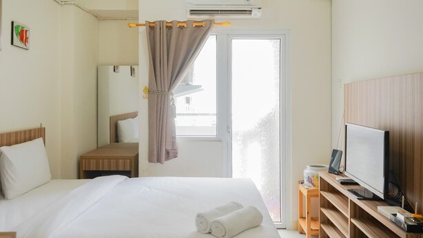 A hotel room with a bed, a TV, a desk, and a window with curtains. - Cozy Studio Green Pramuka Apartment next to Mall (Jakarta)