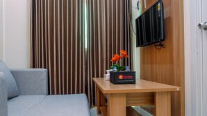 A small room with a grey sofa, a wooden table with a tray table, a brown curtain, and a mounted TV. - Connected to Mall 2BR Apartment at Green Pramuka City (Jakarta)