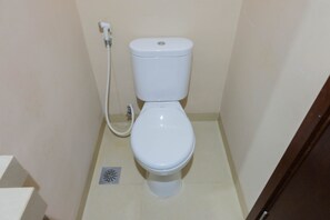 A small, white toilet with a bidet attachment in a narrow bathroom with beige walls and floor.