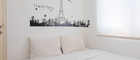 A bedroom with a bed, white bedding, a wall decal of the Eiffel Tower and Paris skyline, and a window with blinds.
