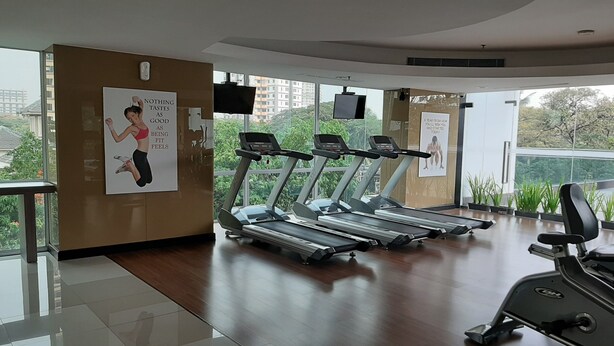 Ein Fitnessstudio mit Laufbändern, einem Hometrainer und einem Rudergerät. Motivationsplakate schmücken die Wände, und große Fenster bieten einen Blick auf die Stadt.