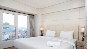 A hotel room with a large bed, two bedside lamps, a view of the city through large windows, and a cityscape visible outside. - Stunning 2BR Loft Apartment at Maqna Residence (Jakarta)