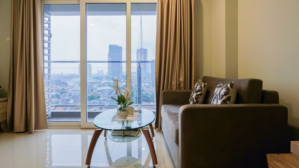 A modern living room with a glass coffee table, a sofa, and a view of a cityscape through large windows. - Modern 2BR Apartment at Maqna Residence (Jakarta)
