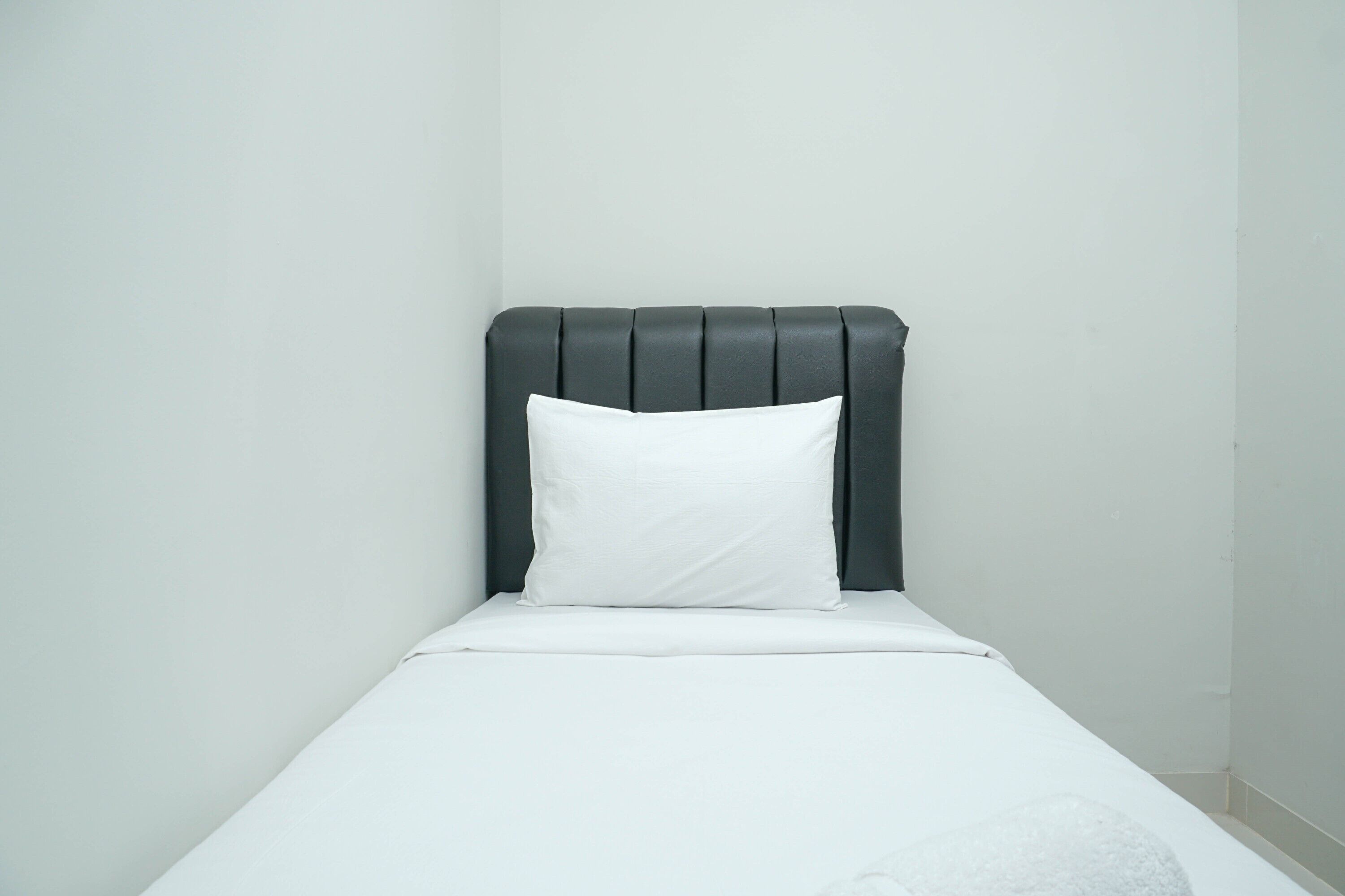 Un letto singolo con una testata scura e un cuscino bianco in una stanza dalle pareti bianche e spoglie.