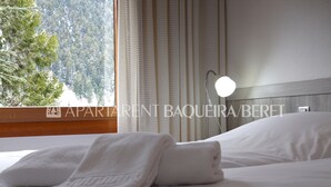 Una cama bien hecha con sábanas blancas y una toalla doblada, una lámpara de noche y una ventana con cortinas.