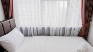 Una cama bien hecha con sábanas blancas, una toalla enrollada y una almohada, frente a cortinas translúcidas y vistas a la ciudad.