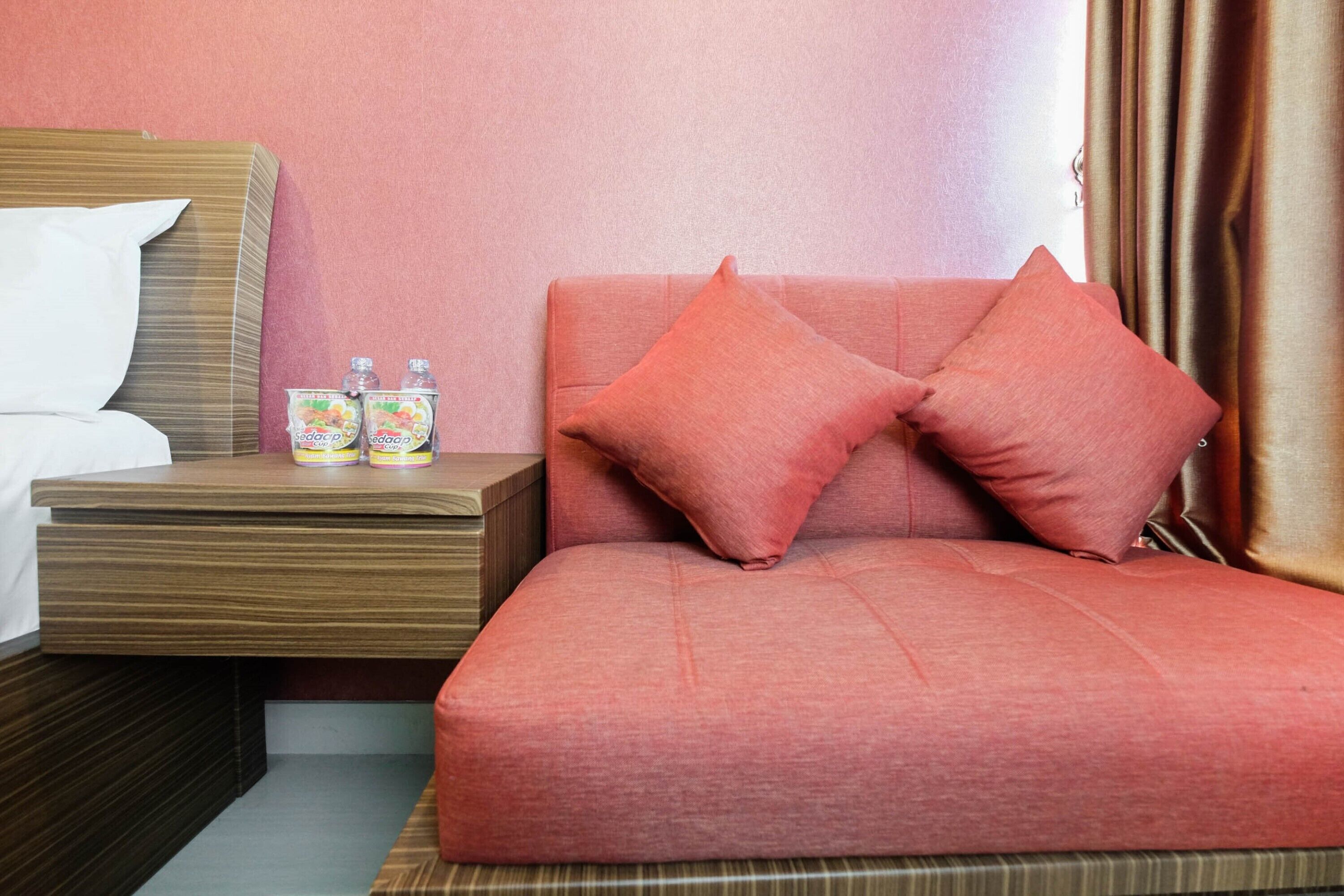 A hotel room with a bed, a wooden nightstand, and a sofa with two pillows. There are two bottles on the nightstand.