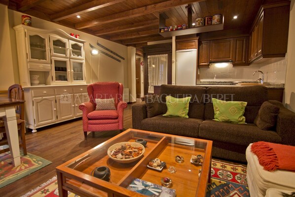 A living room with a red armchair, a brown sofa with green cushions, a coffee table with various items, and a kitchen area with wooden cabinets and a refrigerator.