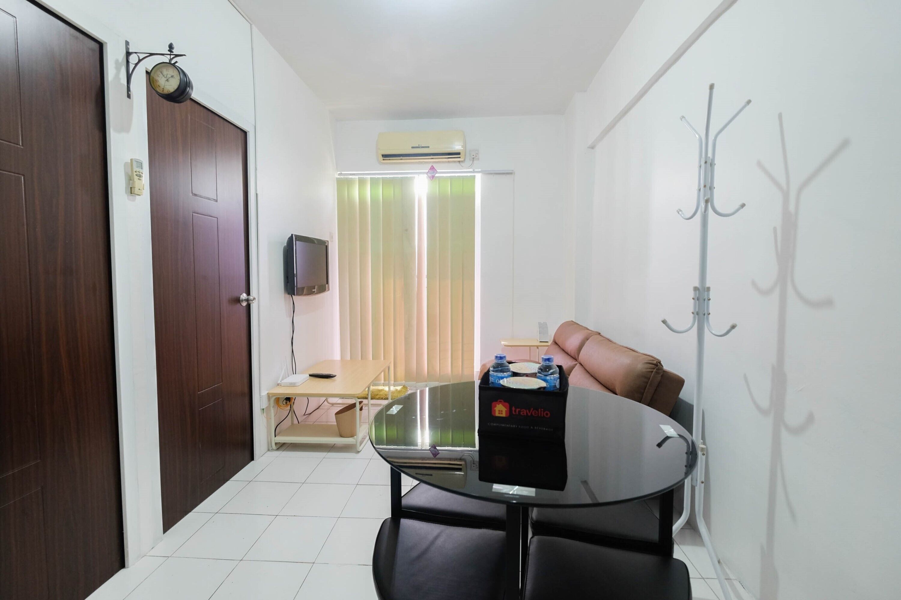 A hotel room with a wooden door, a television on the wall, a window with blinds, a table with a box of toiletries, and a coat rack.