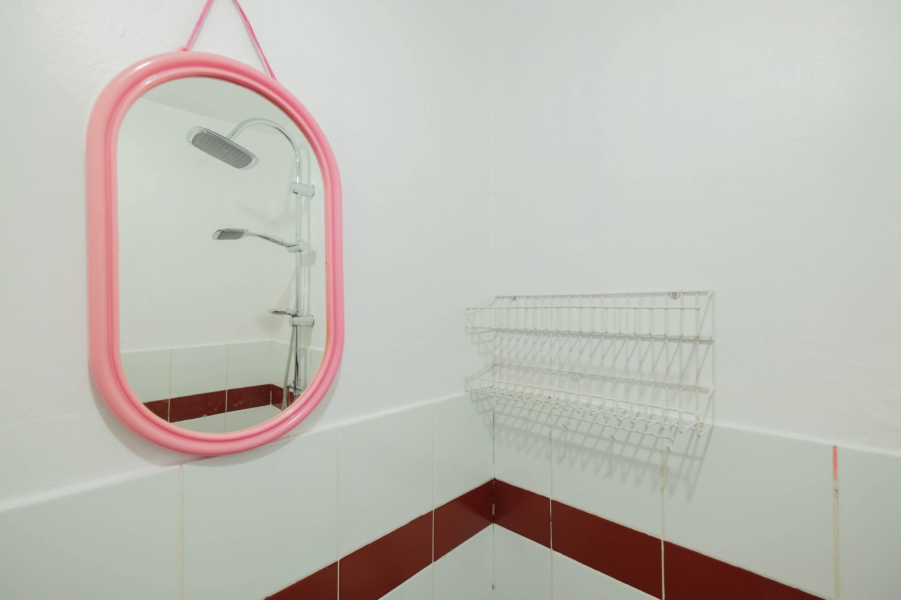 A bathroom with a pink-framed mirror, a shower, and a white tiled wall with a red stripe.