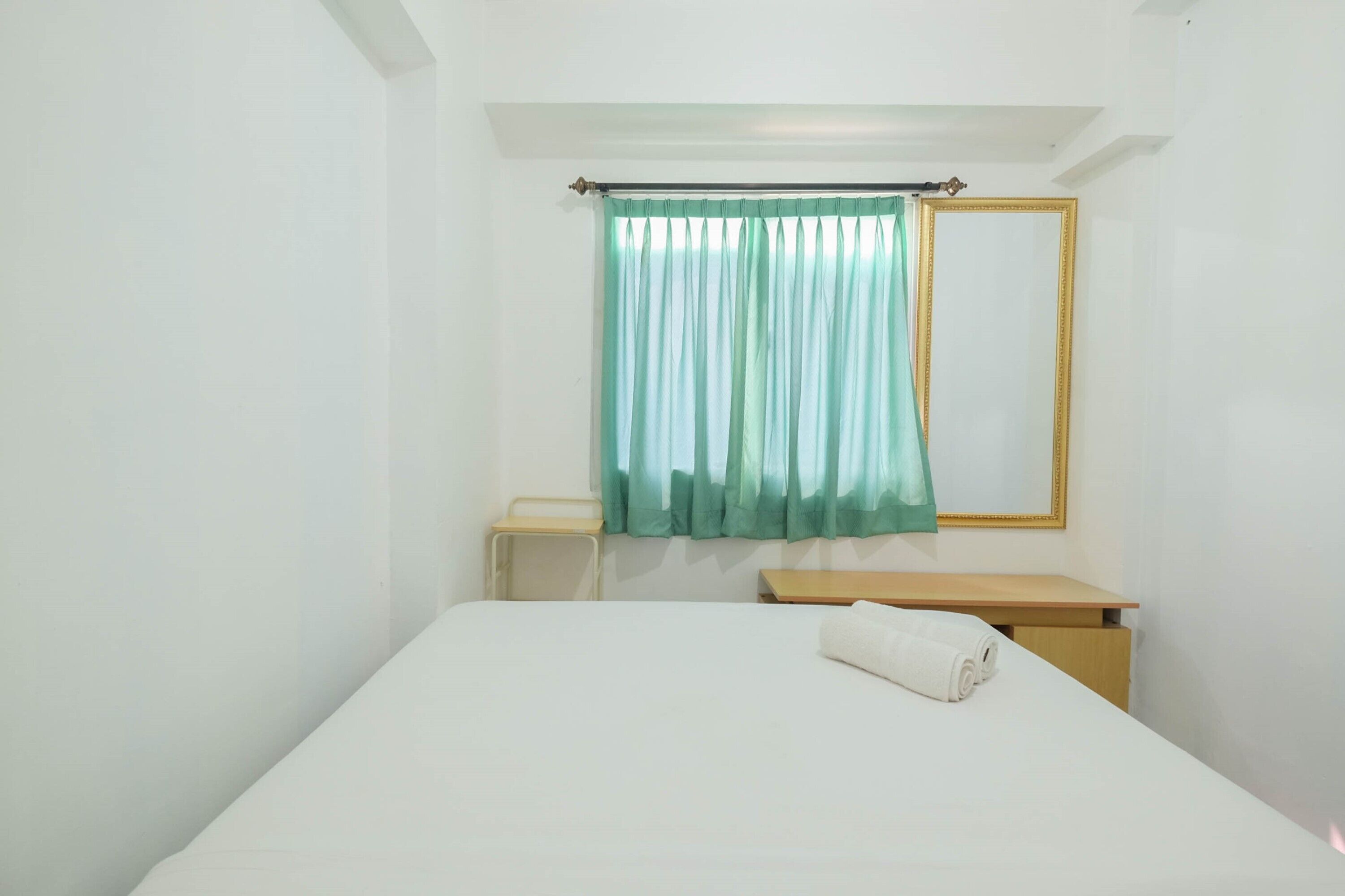 Ein Hotelzimmer mit einem Bett, einem kleinen Nachttisch, einem Holznachtschränkchen, einem Spiegel und einem Fenster mit grünem Vorhang.