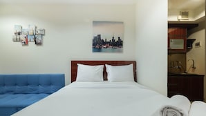 Ett hotellrum med en säng, en blå soffa, ett konstverk som föreställer en stadsbild och ett litet kök.