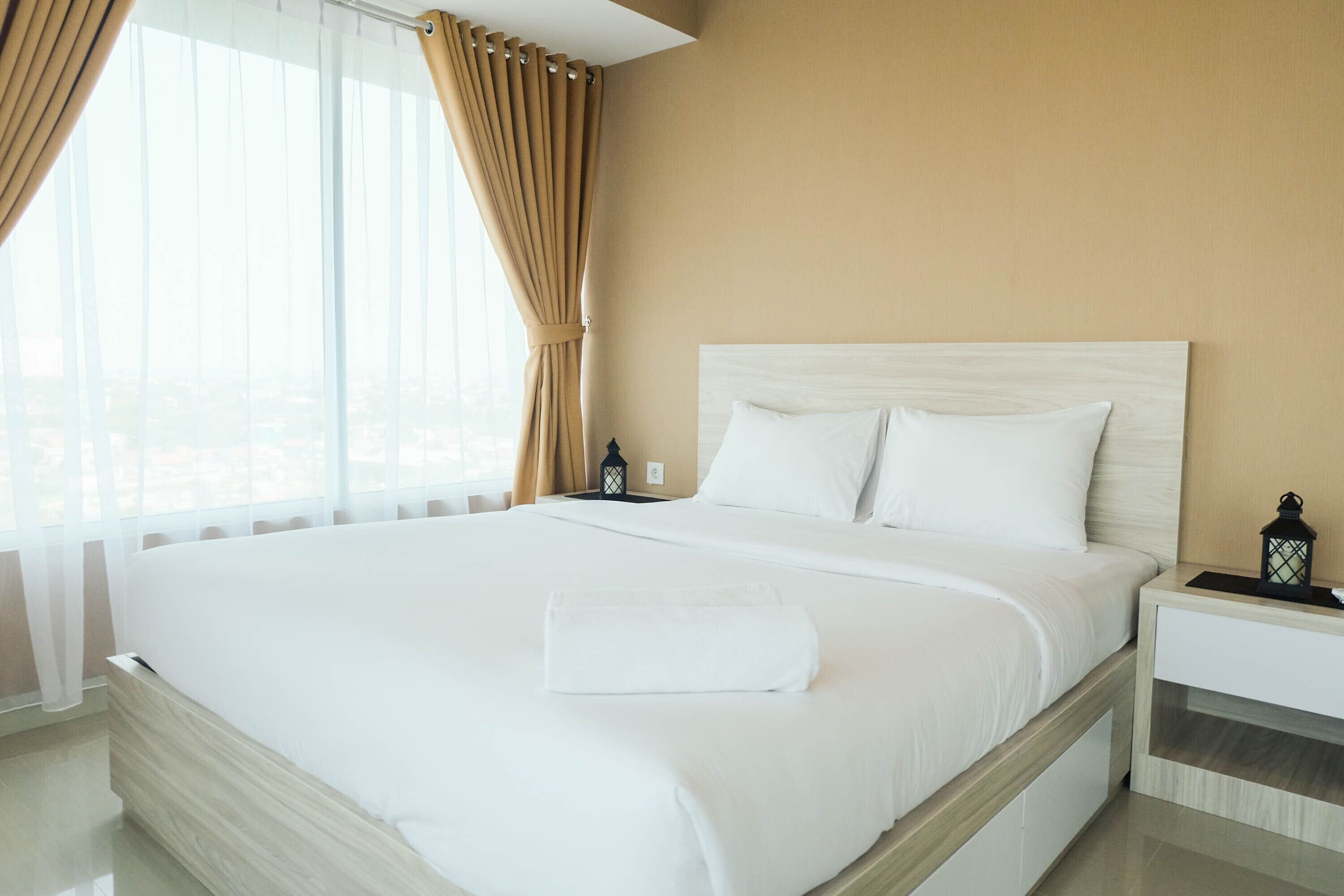 A hotel room with a large bed, a nightstand, a lamp, and a view of the city through the window.