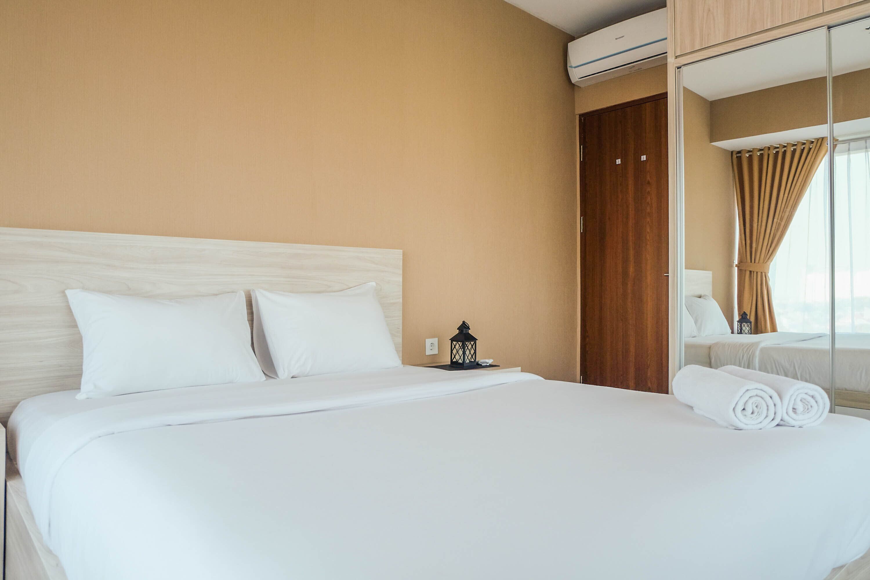 A hotel room with a large bed, a nightstand with a lamp, a mirror, and a view of the outside through a window.