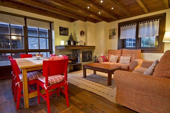 A cozy living room with a fireplace, wooden beams, and a dining area with red chairs.