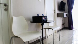 A hotel room with a white chair, a small table with a black bag and water bottles, a TV on a stand, and a door with a handle. - New Furnished 2BR Apartment at Silkwood Residence (Tangerang)