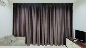 A room with a large window covered by dark curtains, a sofa, a TV on a wooden cabinet, and an air conditioning unit on the wall. - Luxurious and Comfy 2BR Paddington Heights Alam Sutera Apartment (Tangerang)