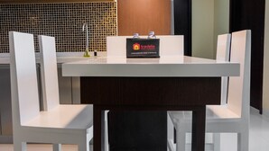 A modern hotel room with a white dining table, two white chairs, and a minibar with a 'Travelio' sign. - Best Price 2BR Apartment at Brooklyn Alam Sutera (South Tangerang)