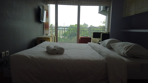 A hotel room with a bed, a TV mounted on the wall, a window with a view of greenery, and a small table with a chair. - Unique Studio Galeri Ciumbuleuit Apartment near Dago (Bandung)