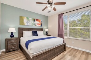 Une chambre à coucher comprenant un lit, deux tables de chevet, un ventilateur de plafond, une fenêtre avec des stores et un tableau accroché au mur.