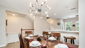 A dining area with a wooden table set for six, surrounded by chairs. A modern chandelier hangs from the ceiling, and a window provides natural light.