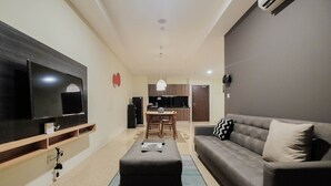 Ein modernes Wohnzimmer mit Flachbildfernseher, grauem Sofa, Holz-TV-Schrank, Essbereich mit Tisch und Stühlen sowie Küche im Hintergrund.