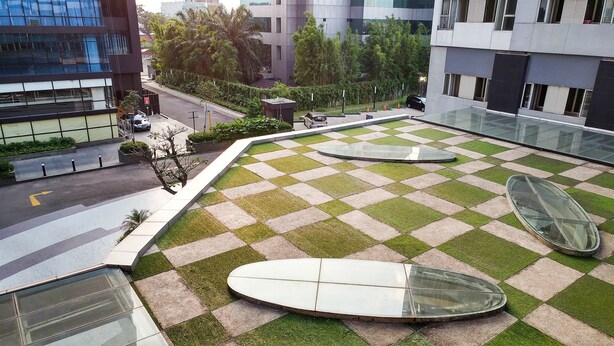 Une terrasse sur le toit, avec un motif à carreaux alternant gazon et carreaux de céramique, une balustrade en verre et quelques voitures garées.