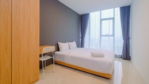 A modern hotel room with a wooden wardrobe, a bed with white linens, a small white desk, and a view of the city through large windows with blue curtains. - Modern and Spacious 2BR L'Avenue Apartment (Jakarta)