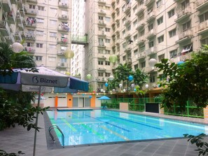 A swimming pool with a tiled deck, surrounded by greenery and a multi-story building with balconies. - Homey and Stylist 1BR Gading Icon Apartment (Jakarta)