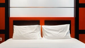 Cama con sábanas blancas y dos almohadas, frente a una pared con un diseño geométrico de paneles negros y naranjas.