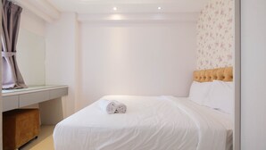 A neatly made bed with white linens and a rolled towel on top, a wooden headboard, and a small wooden bench under a desk with a mirror.