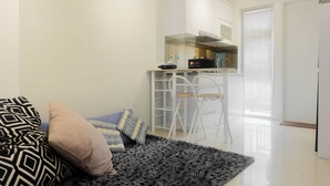 A compact living space with a sofa, a patterned throw, and a dark rug. A kitchenette with white cabinets and a stove is visible in the background. - Stylish and Modern 2BR Bassura City Apartment (Jakarta)