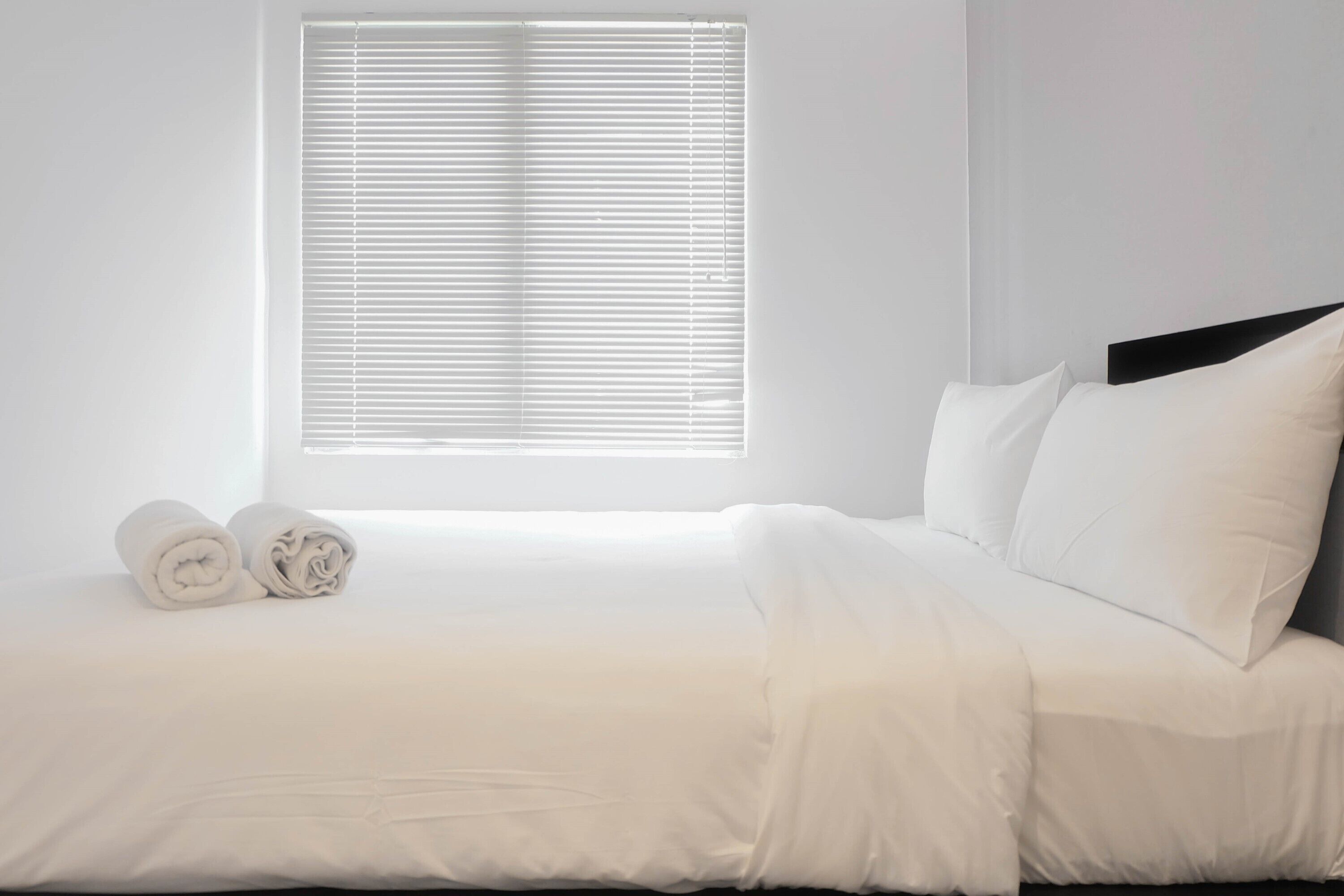 Ein ordentlich bezogenes Bett mit weißen Bettwäsche und Kissen, ein Fenster mit Jalousien und ein zusammengerolltes Handtuch auf dem Boden.