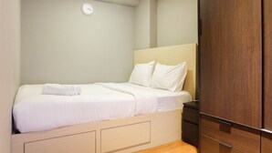 A neatly made bed with white linens and a rolled towel, a wooden headboard, a dark wooden wardrobe, and a wall clock. - Exclusive and Spacious 1BR Apartment at Bassura City (Jakarta)