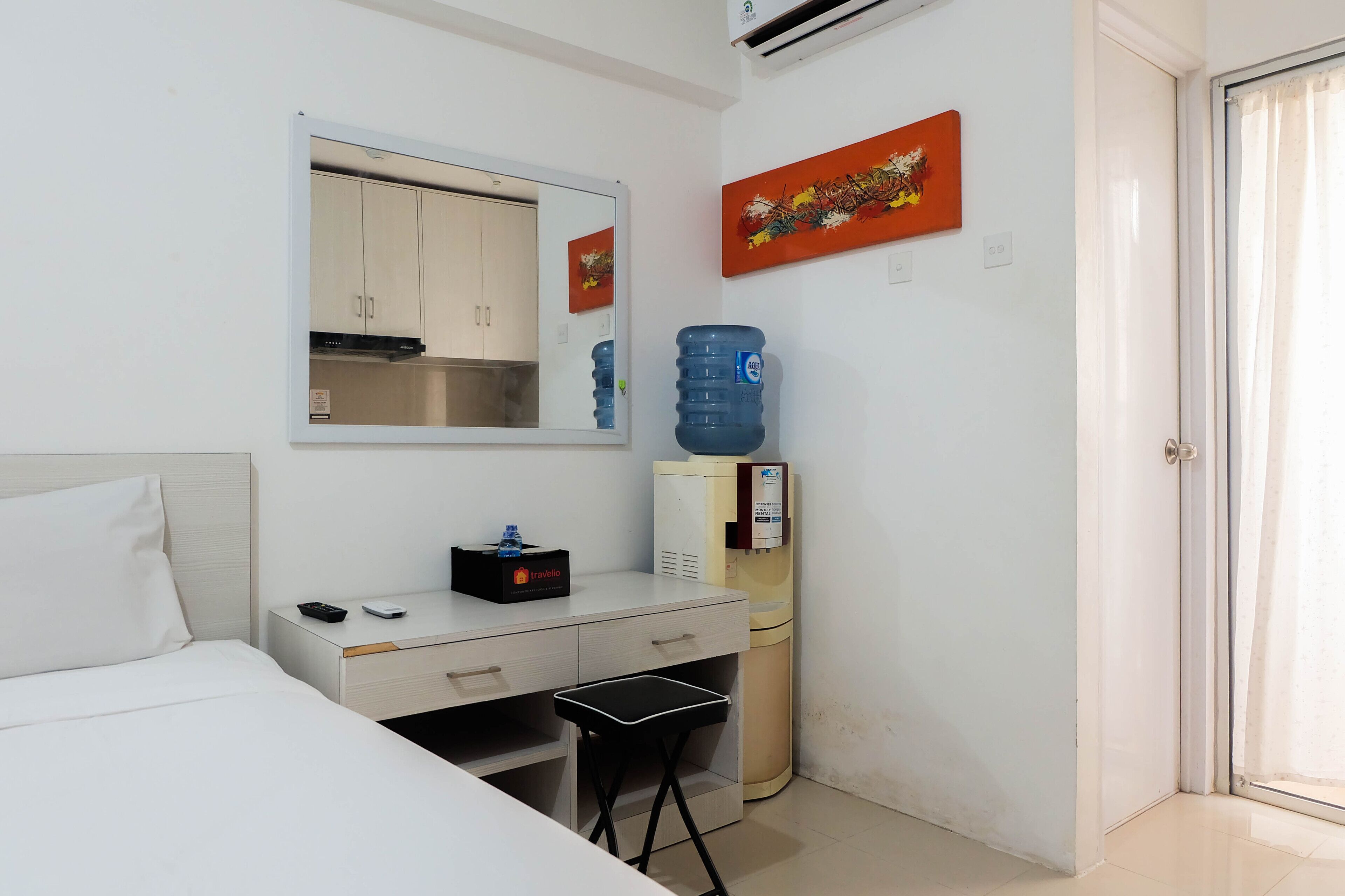 A hotel room with a bed, a desk with a chair, a water dispenser, a mirror, and a door.