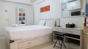 A hotel room with a bed, a desk, a chair, a wardrobe, a mirror, and a water dispenser. - Tidy Studio Apartment at Bassura City near Shopping Mall (Jakarta)