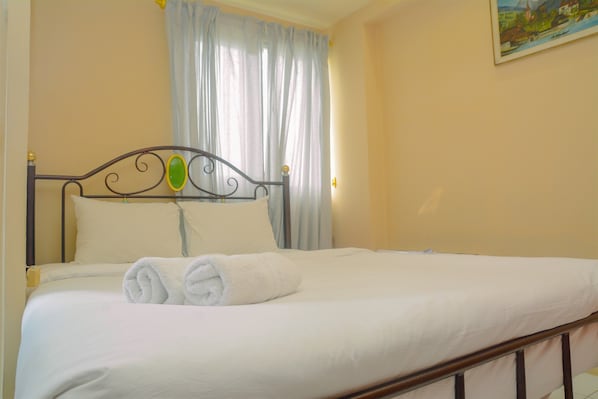 Una cama bien hecha con sábanas blancas y dos toallas enrolladas, un cabecero de metal y una ventana con cortinas traslúcidas.
