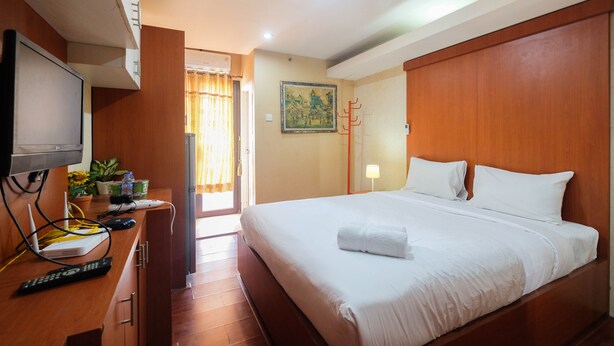 A hotel room with a bed, a desk with a TV, a plant, and a painting on the wall. - Comfy Studio Room Apartment at Kebagusan City (Jakarta)