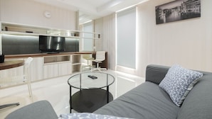 A modern living room with a grey sofa, a glass coffee table, a TV, and a kitchen area with a stainless steel refrigerator and a dining table. - Elegant and Comfy Studio Azalea Suites Apartment Cikarang (Cikarang)