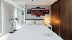 A hotel room with a large bed, a wall-mounted artwork, a bedside table with a lamp, and a standing fan. - Elegant and Comfy Studio Azalea Suites Apartment Cikarang (Cikarang)