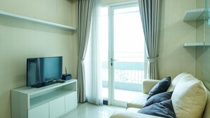A modern living room with a flat-screen TV on a white console, a sofa with cushions, a sliding glass door to a balcony, and a white wall with a shelf. - 2Bedroom Sky Terrace Apartment with Pool View (Jakarta)