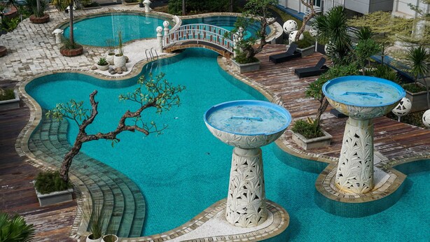 A large, uniquely shaped pool with a bridge, surrounded by tropical plants and decorative pillars. - 2Bedroom Sky Terrace Apartment with Pool View (Jakarta)