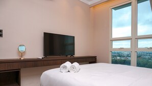 A hotel room with a bed, a TV, a desk with a mirror, and a view of a cityscape through large windows. - Minimalist and Cozy 2BR Citralake Suites Apartment (Jakarta)