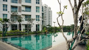 A swimming pool with a wooden deck, surrounded by trees and a modern building in the background. - Minimalist and Cozy 2BR Citralake Suites Apartment (Jakarta)