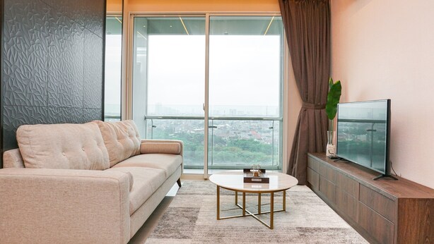 A modern living room with a sofa, a coffee table, a TV on a wooden console, and a large window with a city view. - Minimalist and Cozy 2BR Citralake Suites Apartment (Jakarta)