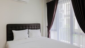 Een net opgemaakt bed met witte lakens en een opgevouwen handdoek, een donker bekleed hoofdbord, en een raam met dunne gordijnen.
