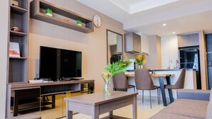 A modern living room with a flat-screen TV, a coffee table, and seating. There is a kitchen area with cabinets and a dining table.